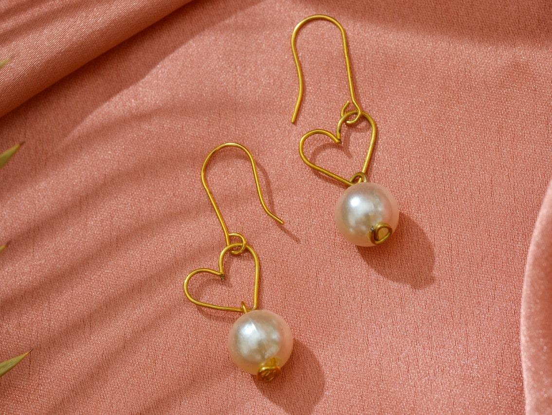 Pink background with yellow gold heart drop earrings with pearls at the bottom