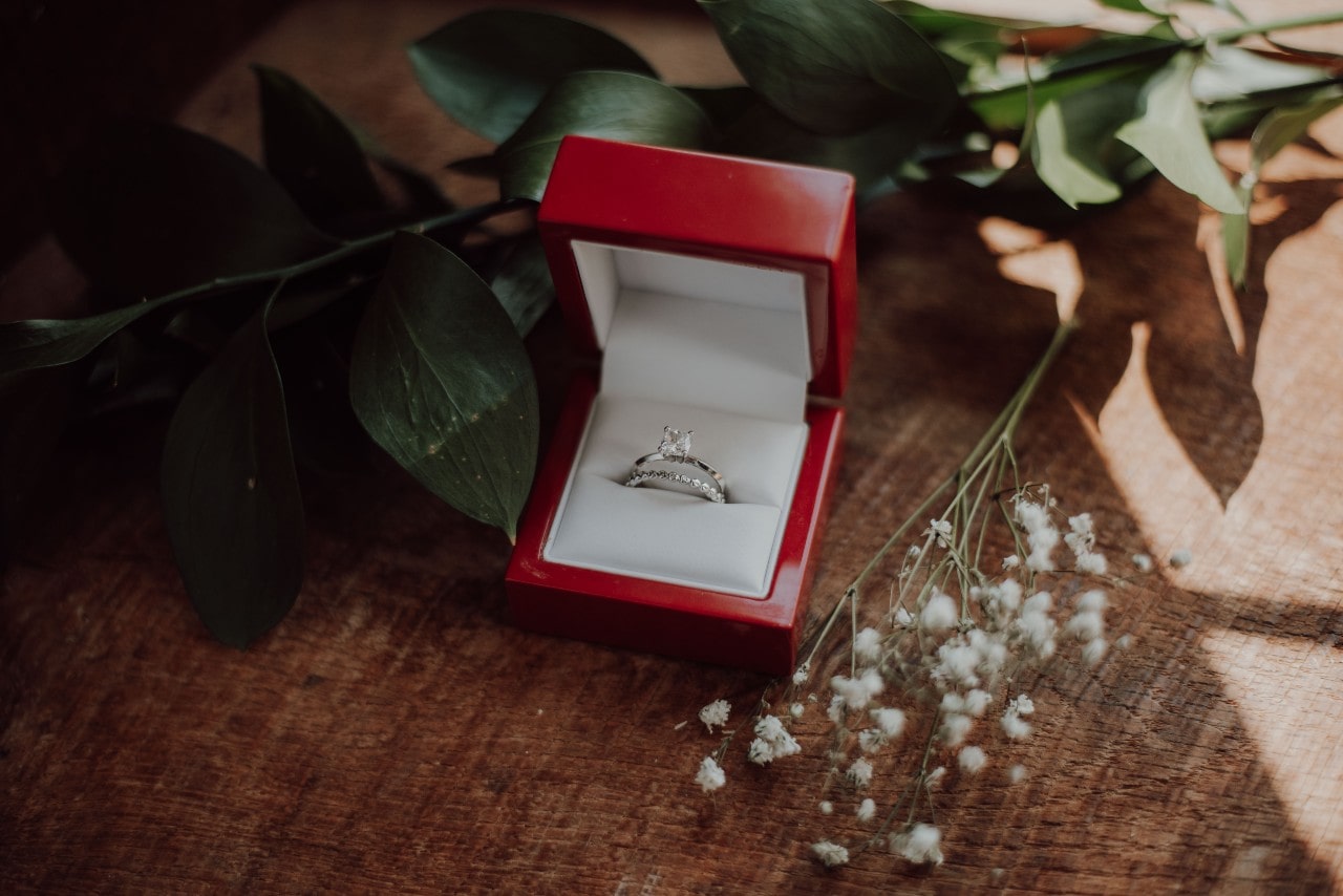 A solitaire princess cut engagement ring in a red ring box with a dainty diamond eternity wedding band