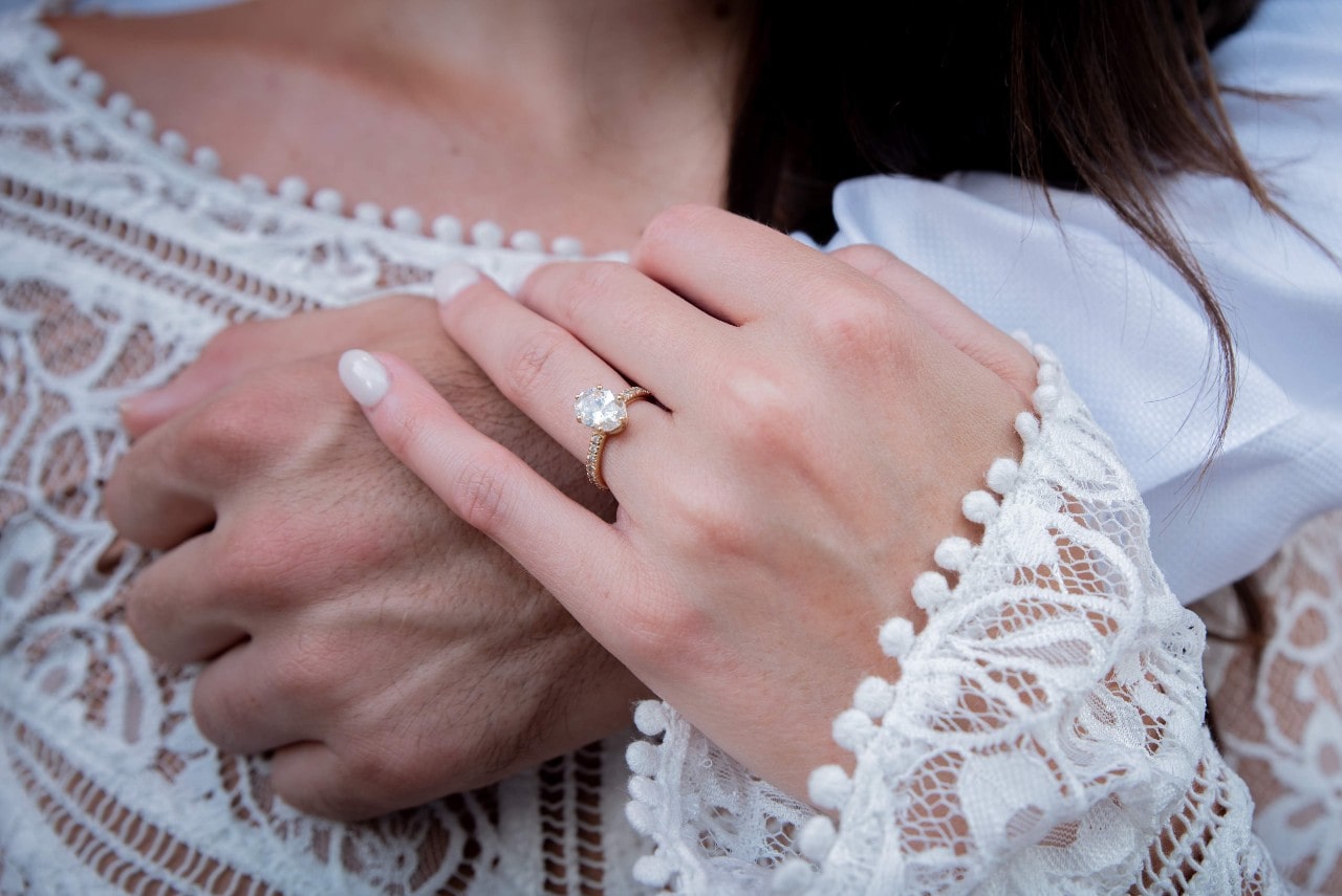 A boho bride resting her hand on her spouse’s showing an oval center stone with diamond side stone band