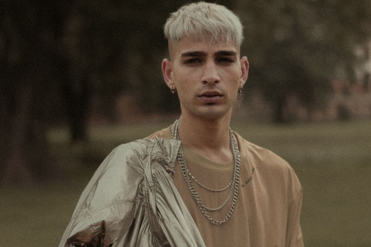 Man looking into the camera wearing a tan t-shirt and three silver chain necklaces