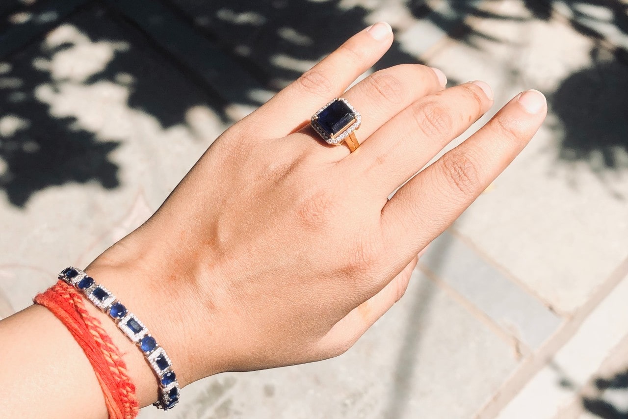 An outstretched hand in the sun wearing a red bracelet, a sapphire line bracelet, and a sapphire ring