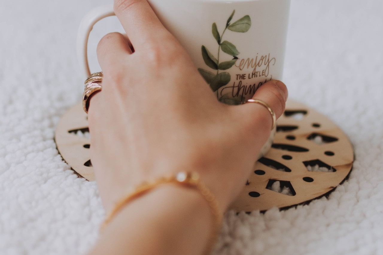 A hand reaching for a white mug wearing a gold bracelet and fashion rings