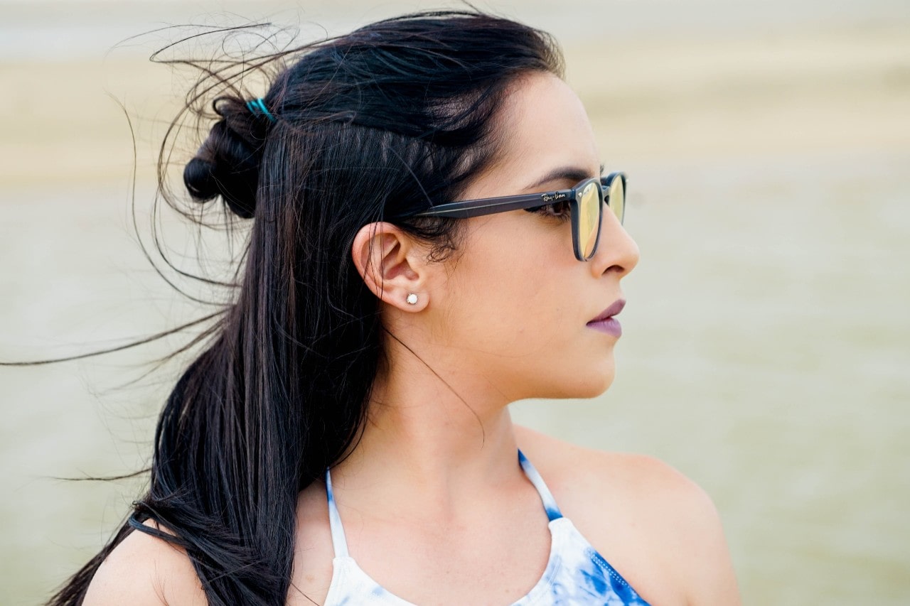 A woman wearing a half-updo wears stud earrings and sunglasses while walking on a beach