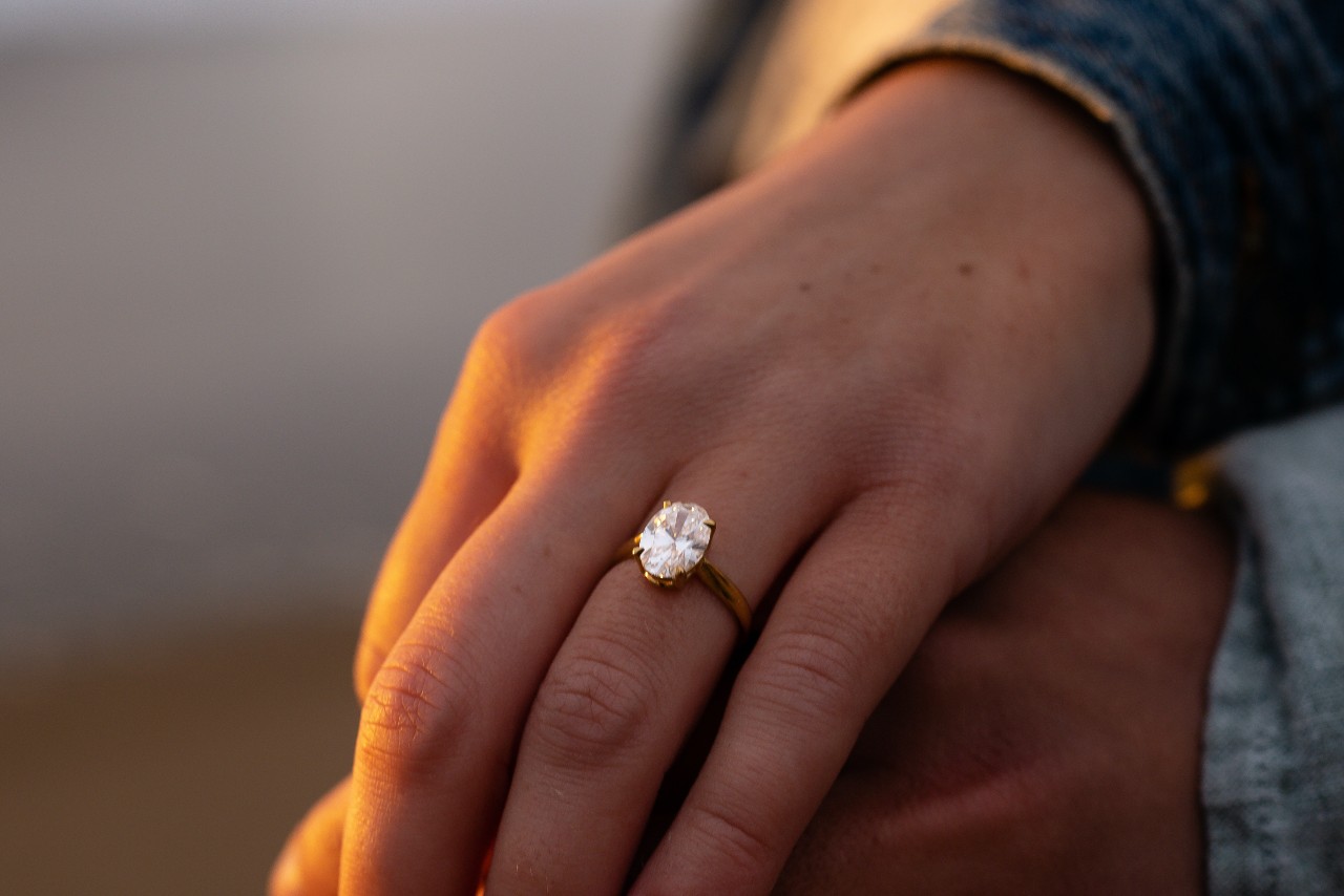 close up of an oval center stone on a yellow gold band on a woman’s hand holding a man’s hand during a sunset