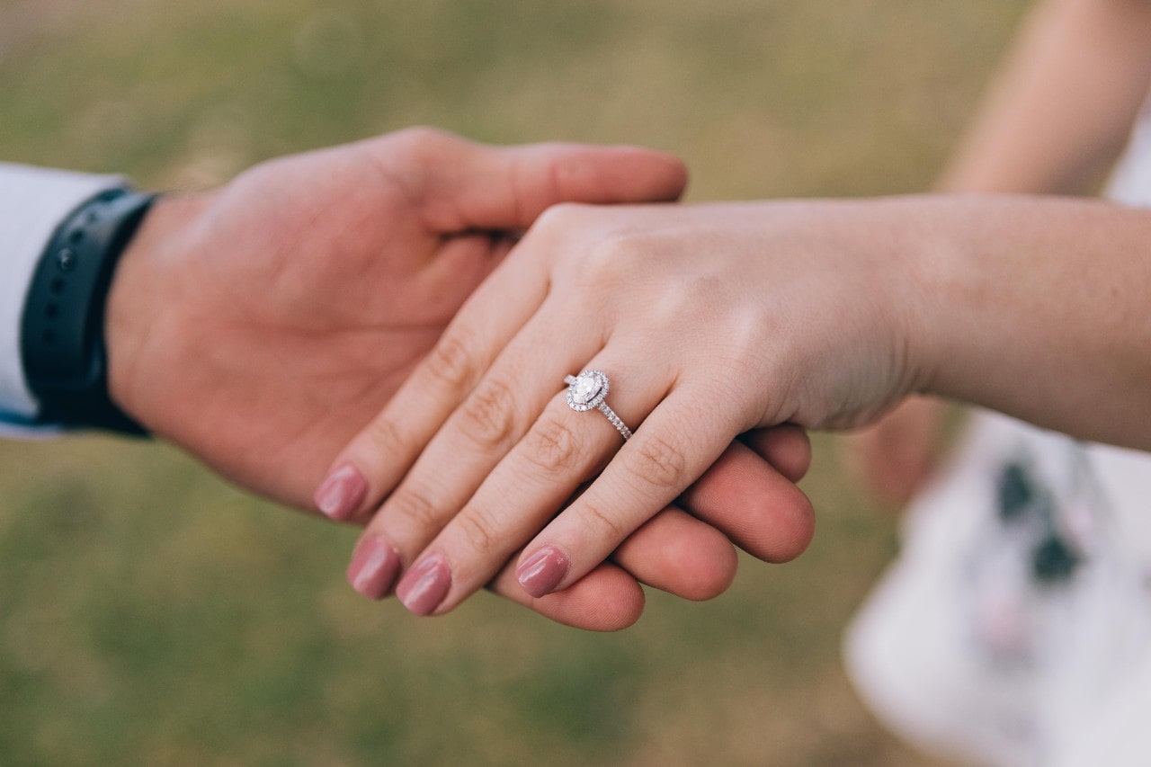 diamond halo engagement ring with side stones along the band, as shown on a woman’s hand being held by her love