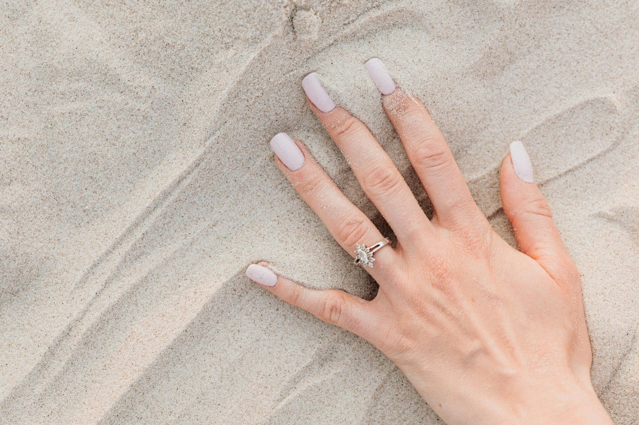 A woman displaying her marquise with halo engagement ring on her hand on top of the sand at the beach.