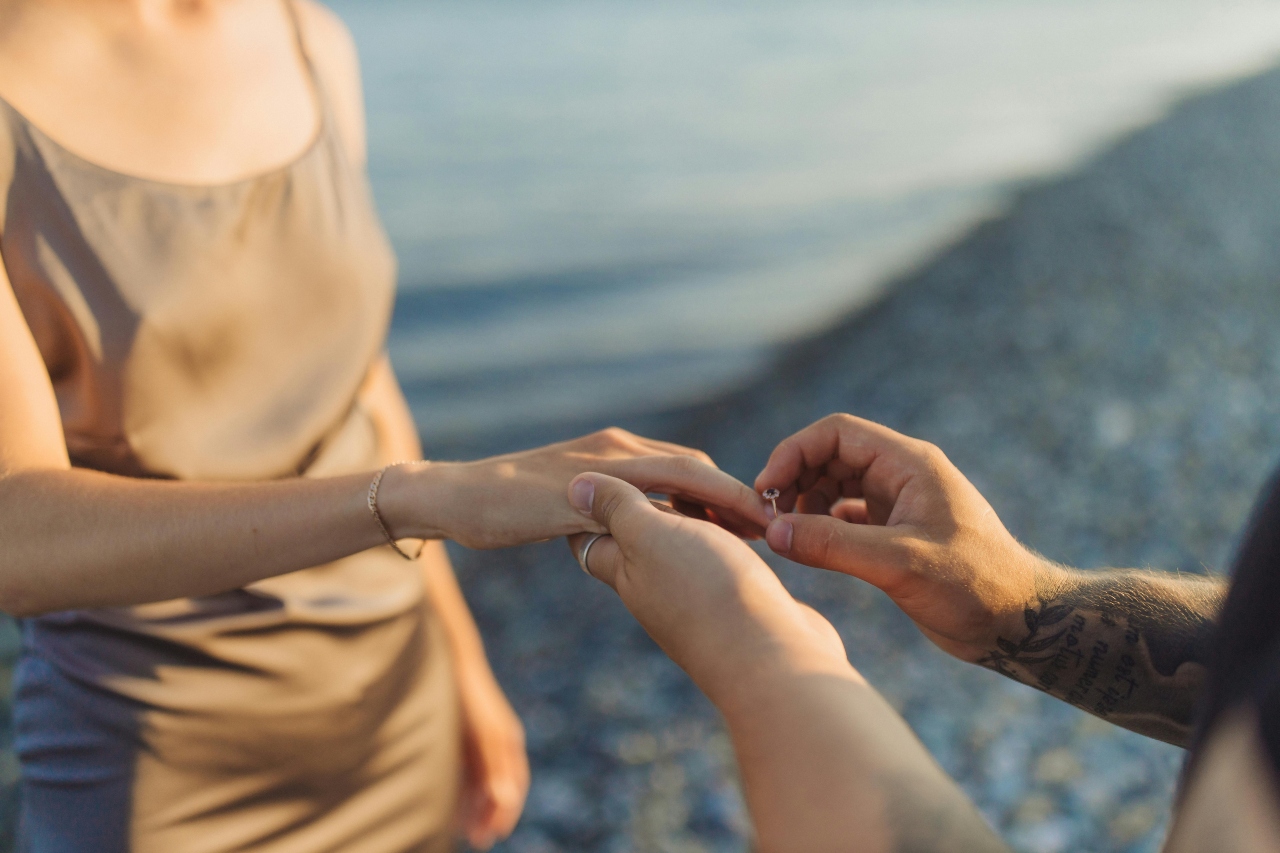 A man putting a sapphire center stone engagement ring with a diamond halo on a woman’s finger after his beachside proposal.