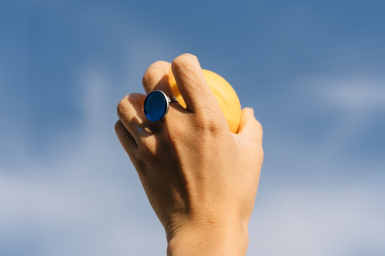 A woman holds an orange up to the sky while wearing a chunky sapphire fashion ring.