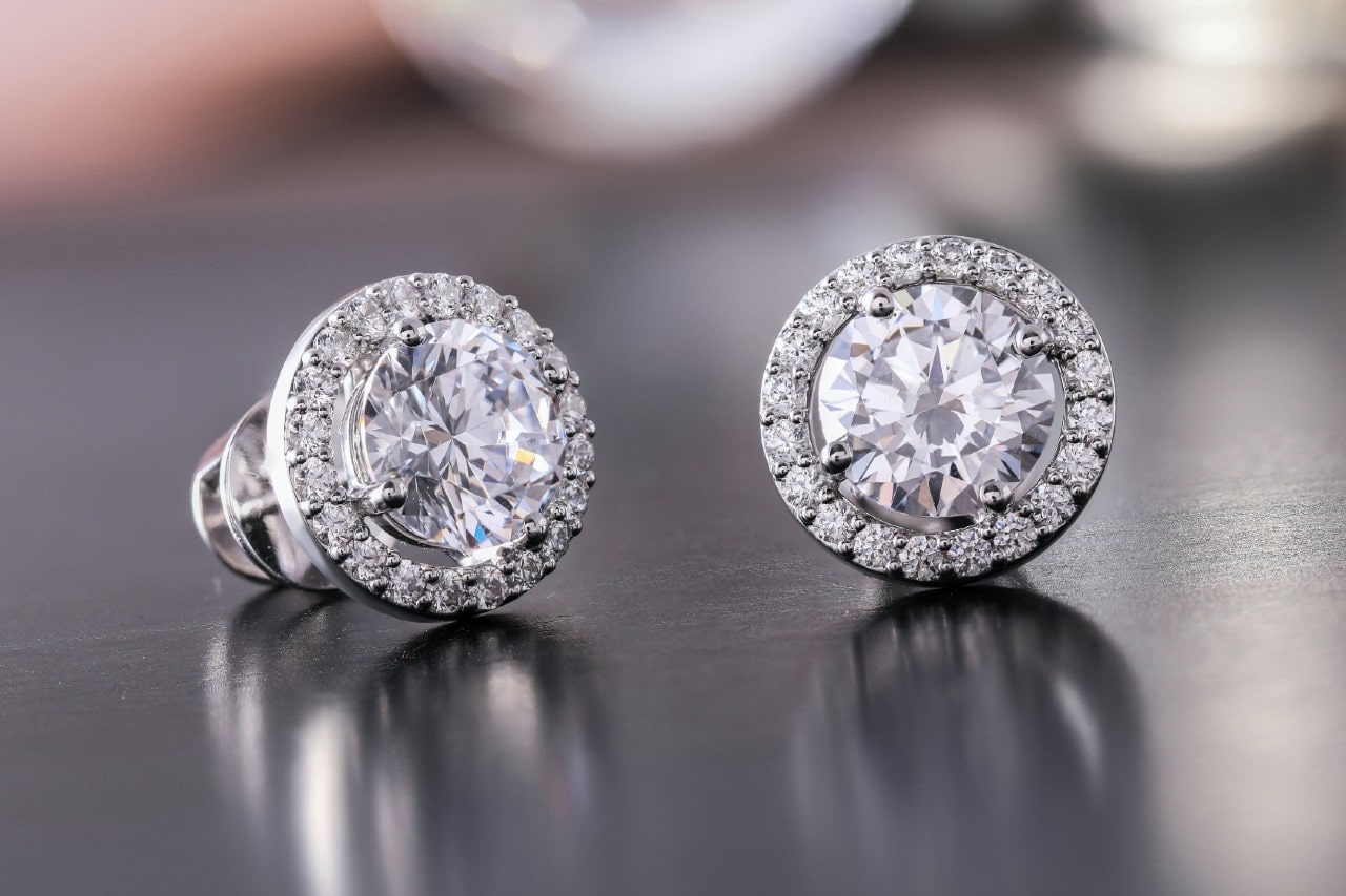 a pair of white gold diamond stud earrings in a halo setting on a reflective surface