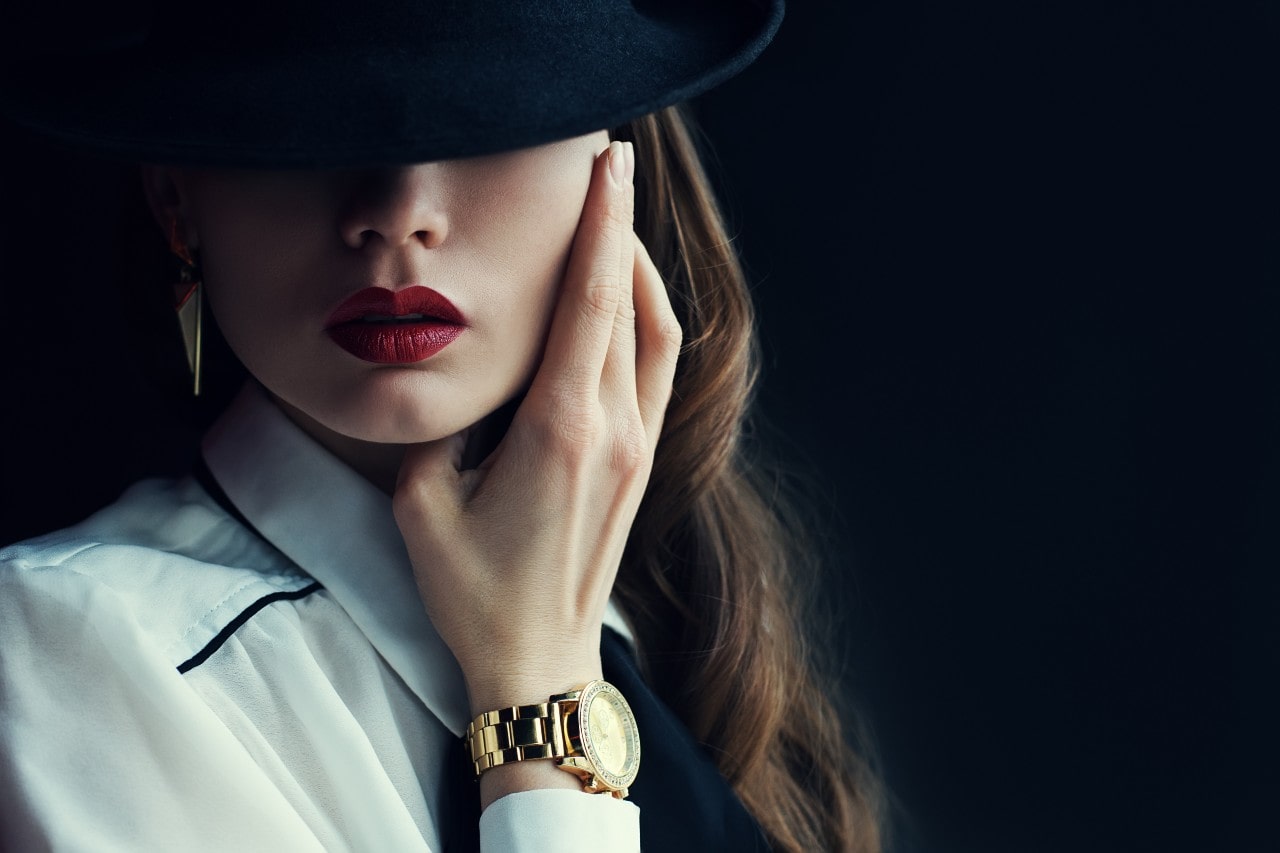 A close-up of a stylish woman wearing a gold watch, her eyes obscured by her hat.