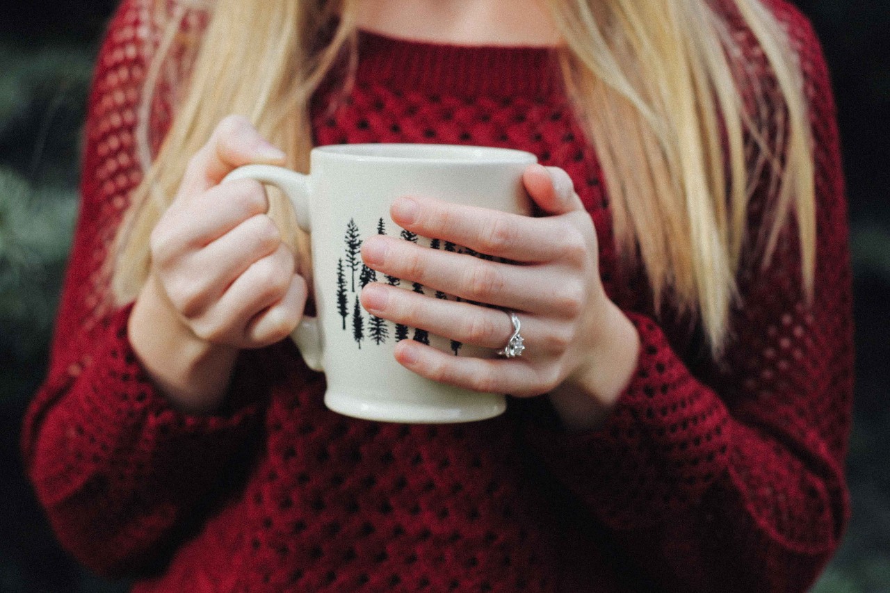 A woman in a red sweater sips coffee in a Christmas mug while wearing her engagement ring.