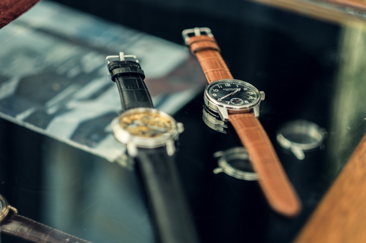 Two elegant wristwatches with leather straps are displayed on a reflective glass surface, with a blurred background.