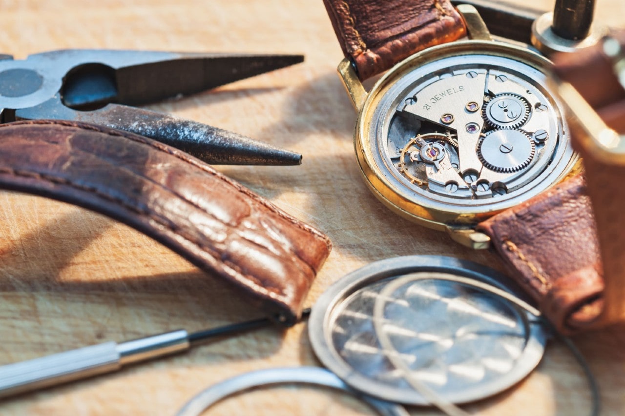 A close-up shot of a vintage watch with its back cover removed and mechanism exposed, next to watch repair tools.
