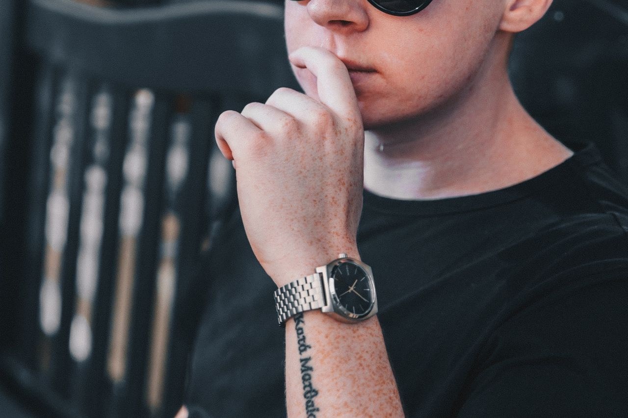 A young man in sunglasses and a black shirt rests his chin on his hand, a stainless steel watch on his wrist.