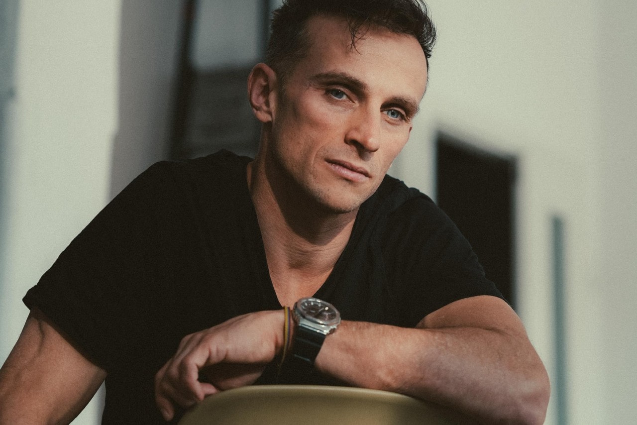 A close-up of a comfortably dressed man in a black t-shirt and sports watch sitting backwards in a chair.