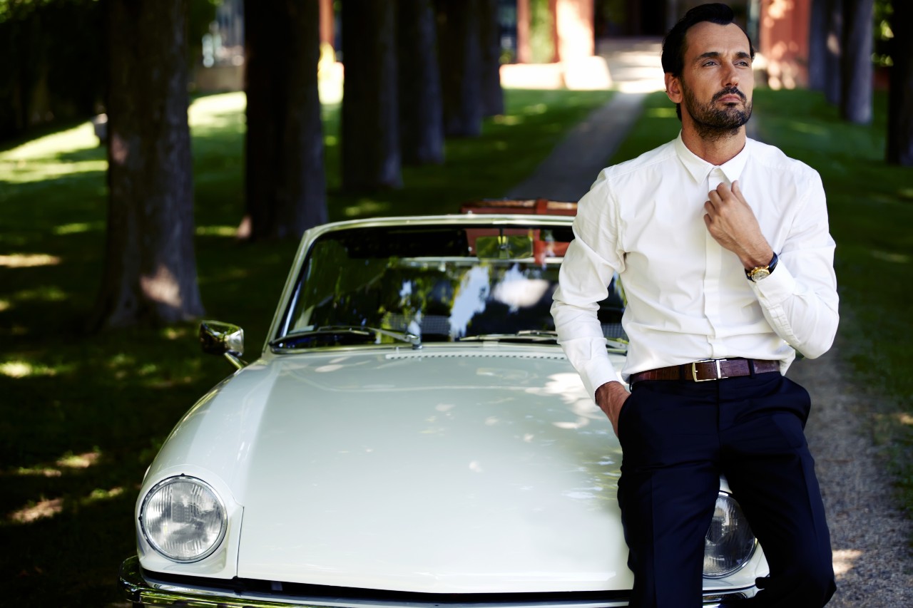 A man leans on a classic car, fixing his collar while wearing a watch.