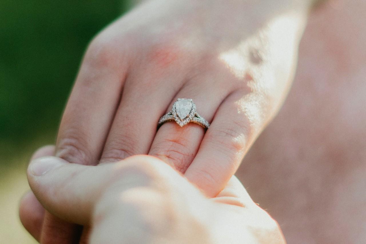 two hands holding each other, one of which is adorned with a halo pear shape engagement ring