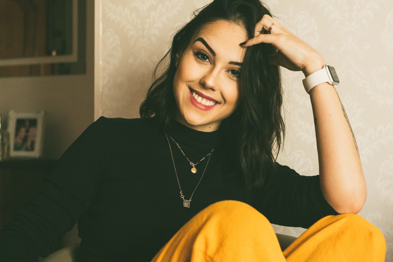 a woman smiling leaning her head on her hand and wearing two gold necklaces