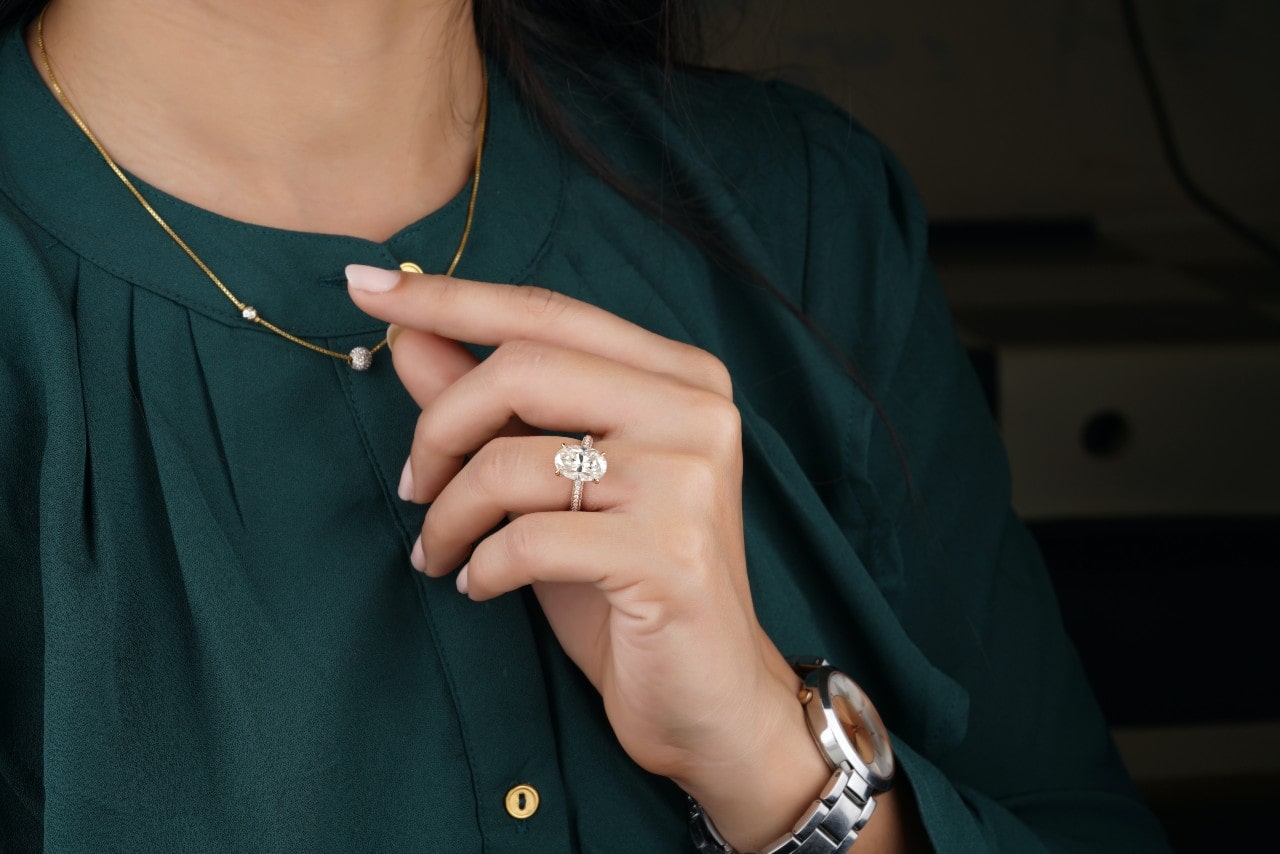 A woman touches her necklace, showing her oval cut side stone engagement ring.