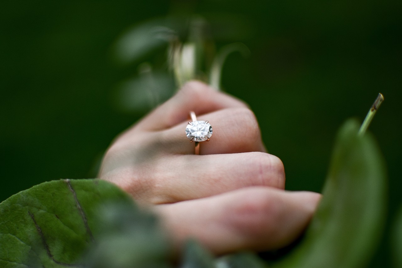 A closeup of a gold solitaire oval cut ring on a woman’s hand as she grasps a branch.