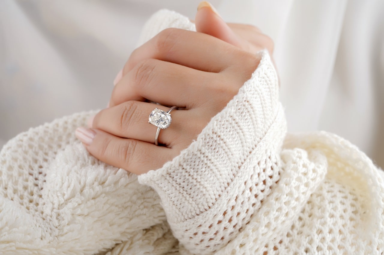 A closeup of a platinum oval-cut diamond solitaire engagement ring on a sweatered woman’s hands.