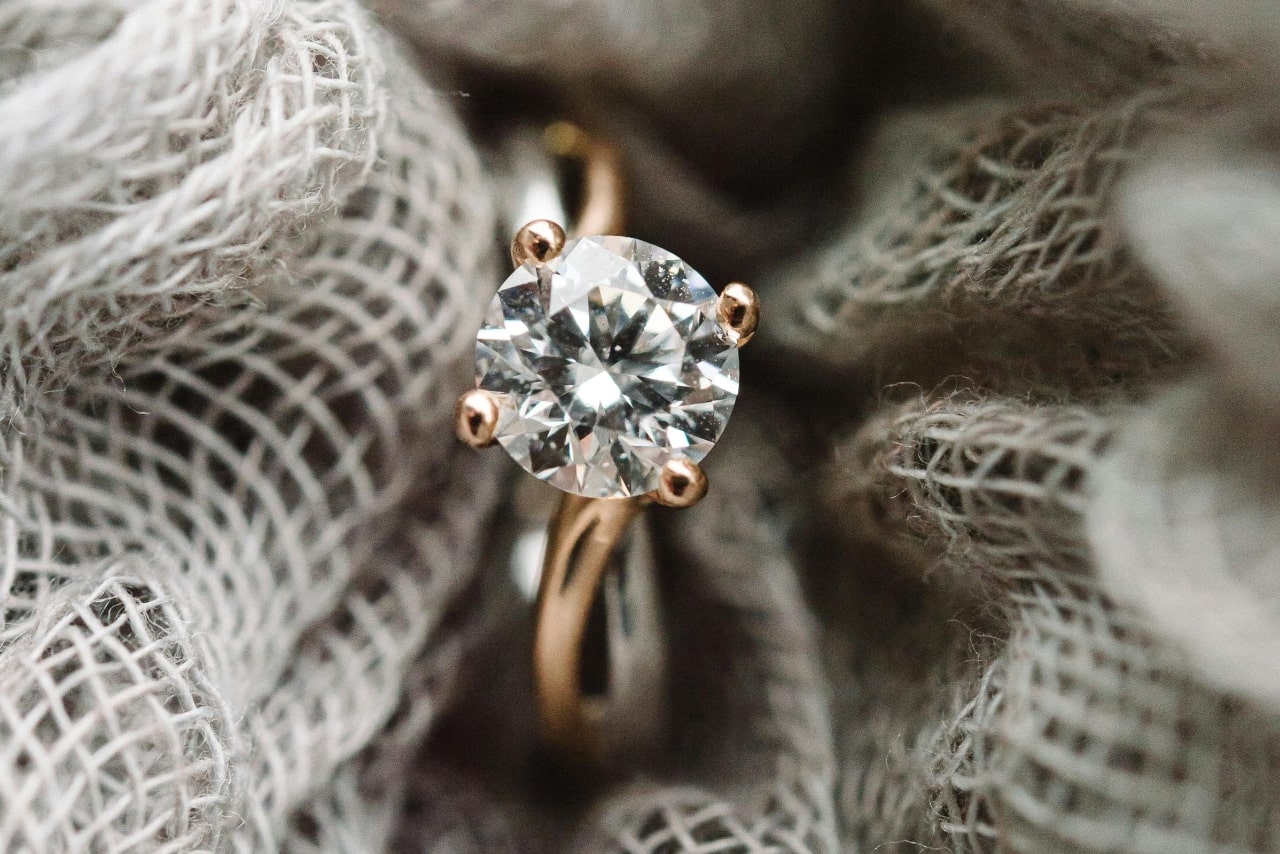 A close-up of a gold engagement ring with a round-cut diamond solitaire set against a textured fabric background. A close-up of a gold engagement ring with a round-cut diamond solitaire set against a textured fabric background.