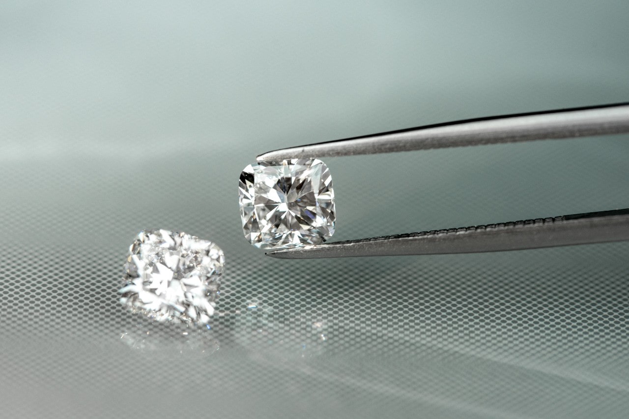 Two cushion-cut diamonds, one held by tweezers, against a reflective surface. Two cushion-cut diamonds, one held by tweezers, against a reflective surface.