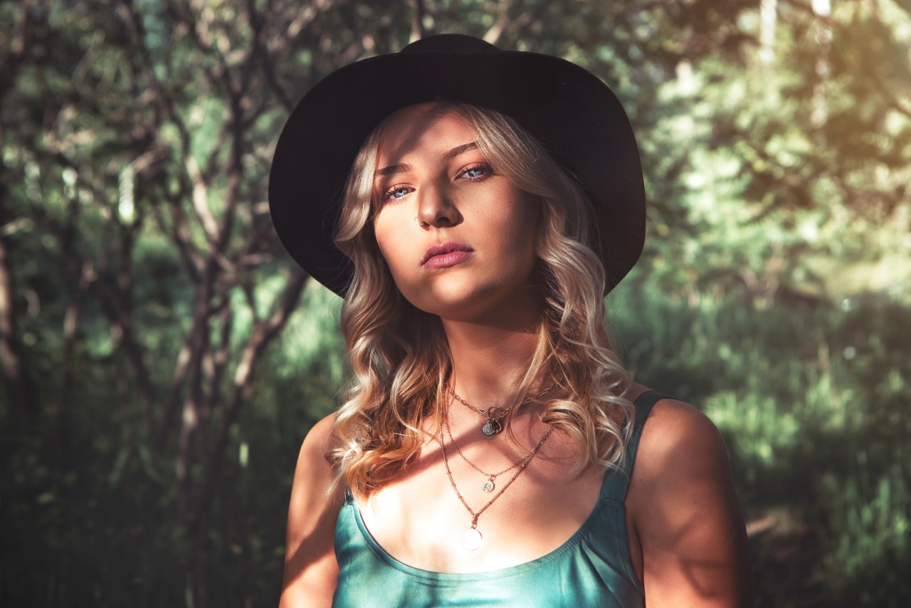 A woman near trees wearing a hat and multiple gold necklaces