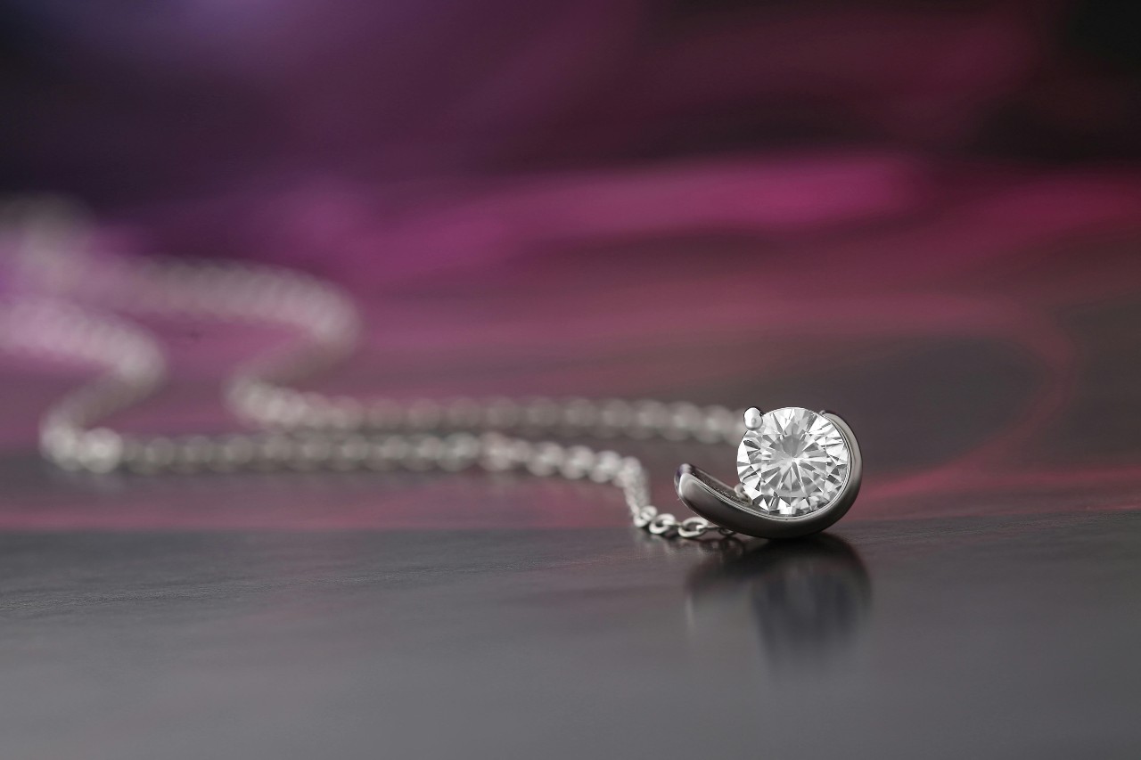 A delicate silver necklace with a round-cut diamond held in a crescent-shaped setting, resting on a dark reflective surface with a soft purple background. A delicate silver necklace with a round-cut diamond held in a crescent-shaped setting, resting on a dark reflective surface with a soft purple background.