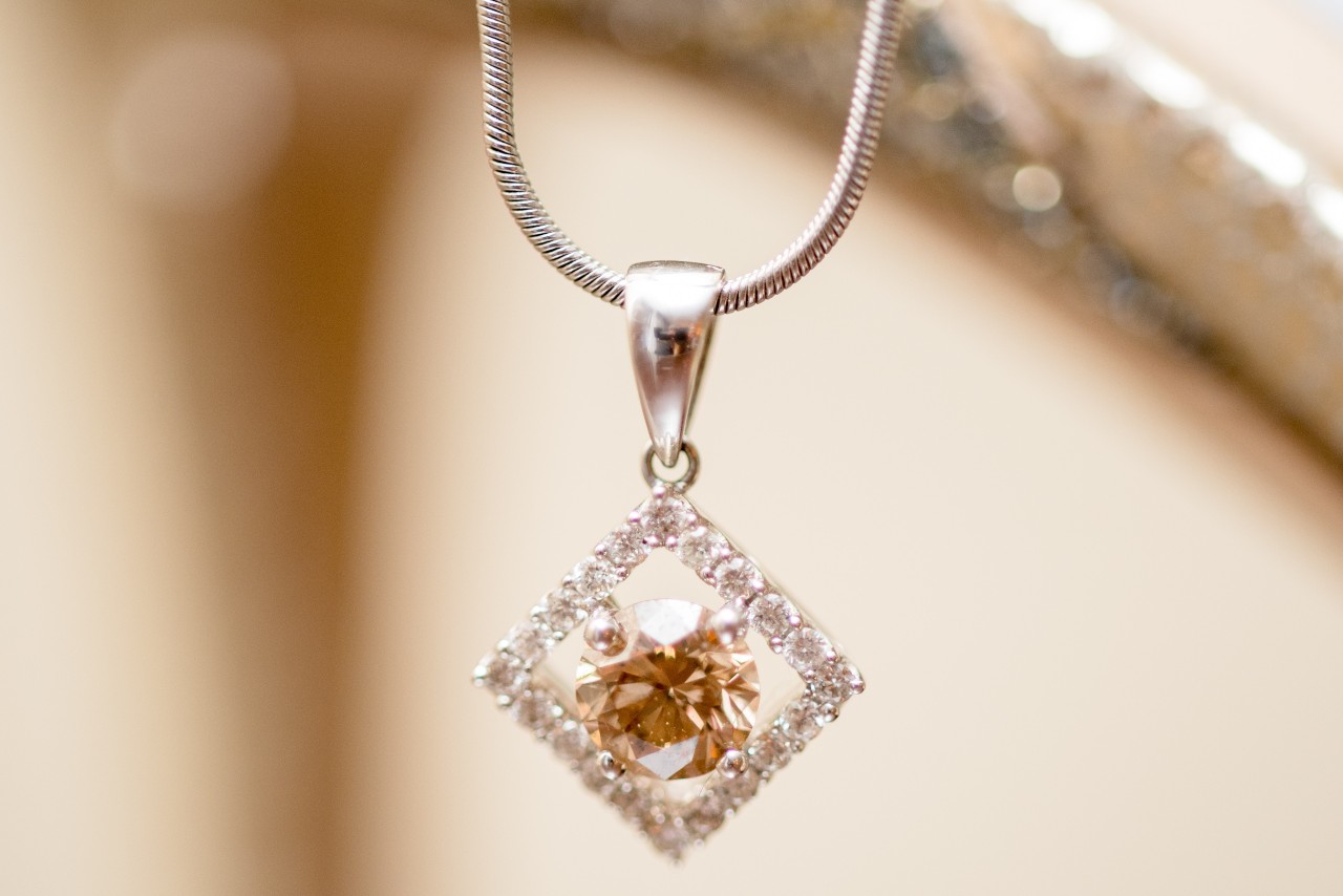 A close-up of a diamond-shaped pendant with a brown gemstone center, surrounded by smaller clear stones, hanging from a silver chain. A close-up of a diamond-shaped pendant with a brown gemstone center, surrounded by smaller clear stones, hanging from a silver chain.