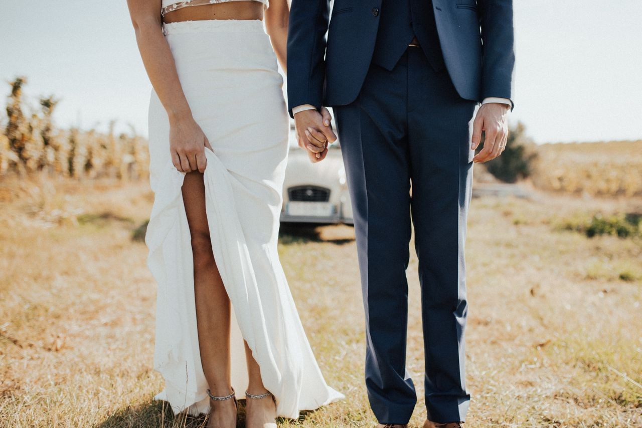 A newly-wed couple holds hands in front of their getaway car in a field.